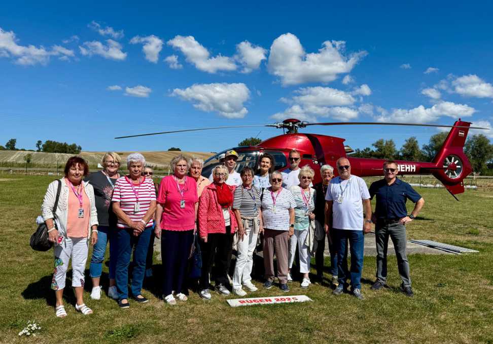 Gruppe von Erwachsenen steht vor einem roten Helikopter auf einer Wiese unter blauem Himmel mit Wolken, Schild „Rügen 2025“ liegt am Boden davor.