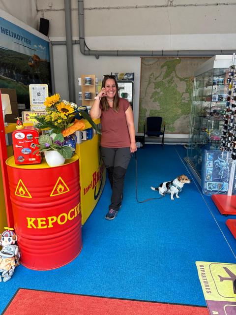 Eine Frau steht lächelnd mit einem kleinen Hund an der Leine an einem gelben Empfangstresen mit der Aufschrift „Insel Rügen“. Daneben steht eine rote Tonne mit Blumen und Karten. Im Hintergrund ist eine große Karte von Rügen und eine Glasvitrine mit Souvenirs zu sehen.