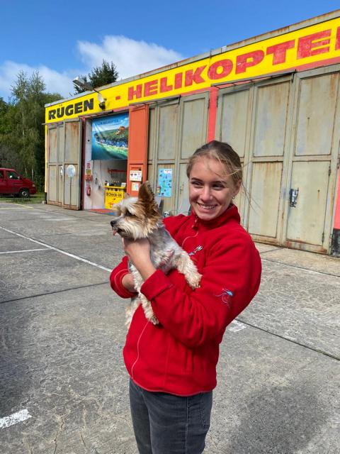 Eine lächelnde junge Frau in einer roten Jacke hält einen kleinen Hund, vor einem Gebäude mit der Aufschrift "Rügen - Helikopter".